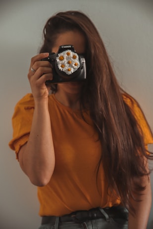 A person with long brown hair is holding a camera up to their face, obscuring it. The camera lens is decorated with white daisies. The person is wearing an orange shirt and standing against a light-colored background.