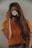 A person with long brown hair is holding a camera up to their face, obscuring it. The camera lens is decorated with white daisies. The person is wearing an orange shirt and standing against a light-colored background.