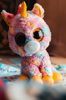 A colorful plush toy with vibrant patterns of pink, blue, and orange, featuring a large pair of shiny eyes and a cute expression. The toy has small plush ears and a prominent yellow horn, suggesting it is an imaginative creature, possibly a unicorn.