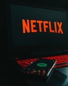 A laptop displays the Netflix logo on its screen while sitting on a desk. The keyboard is illuminated with red backlighting, creating a vivid contrast. In the foreground, a smartphone is visible with a blurry reflection of the Netflix logo.