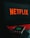 A laptop displays the Netflix logo on its screen while sitting on a desk. The keyboard is illuminated with red backlighting, creating a vivid contrast. In the foreground, a smartphone is visible with a blurry reflection of the Netflix logo.