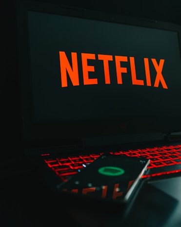 A laptop displays the Netflix logo on its screen while sitting on a desk. The keyboard is illuminated with red backlighting, creating a vivid contrast. In the foreground, a smartphone is visible with a blurry reflection of the Netflix logo.