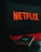 A laptop displays the Netflix logo on its screen while sitting on a desk. The keyboard is illuminated with red backlighting, creating a vivid contrast. In the foreground, a smartphone is visible with a blurry reflection of the Netflix logo.