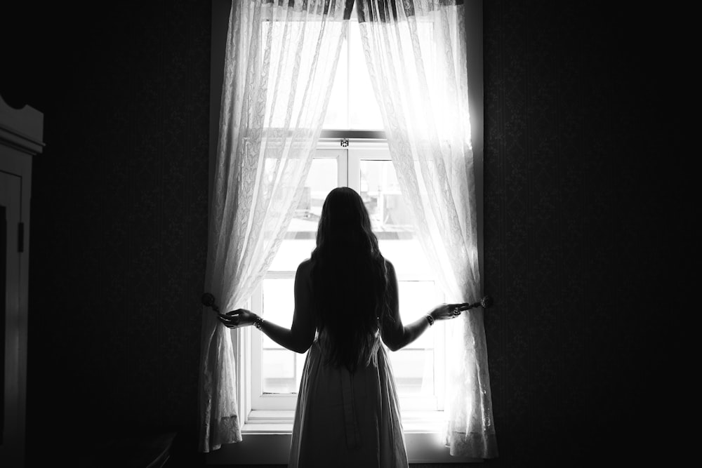 grayscale photo of woman in dress standing near window