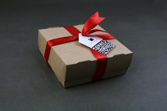 A brown cardboard gift box is wrapped with a vibrant red ribbon and tied in a bow. A small tag with a geometric black and white pattern is attached to the ribbon.