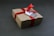 A brown cardboard gift box is wrapped with a vibrant red ribbon and tied in a bow. A small tag with a geometric black and white pattern is attached to the ribbon.