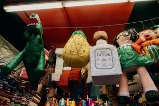 A collection of colorful piñatas hanging from a ceiling in a market stall. There is a green dinosaur, a pineapple, and another shaped like a bottle. Other characters include a person with glasses and bright clothing.