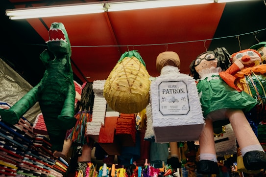 A collection of colorful piñatas hanging from a ceiling in a market stall. There is a green dinosaur, a pineapple, and another shaped like a bottle. Other characters include a person with glasses and bright clothing.