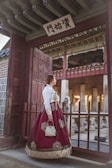A person wearing a traditional Korean hanbok stands in an ancient architectural setting, characterized by wooden gates and intricate roof designs. The hanbok is embellished with gold patterns and the person carries a small handbag.