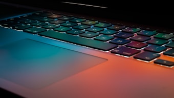 A laptop keyboard is illuminated by colorful ambient lighting reflecting off its surface. The image focuses on the keys and part of the trackpad, capturing the sleek design and modern aesthetic.