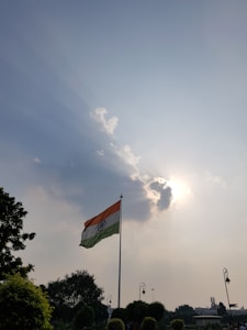 A tall flagpole with the Indian national flag stands prominently against a backdrop of a partly cloudy sky. Sunlight filters through the clouds, creating a dramatic effect with light rays stretching outward. Trees and lamps are visible at the bottom, framing the scene.