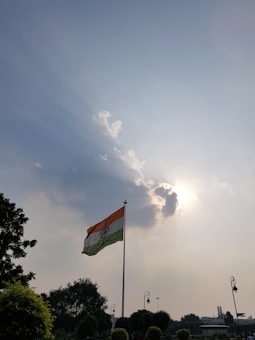 A tall flagpole with the Indian national flag stands prominently against a backdrop of a partly cloudy sky. Sunlight filters through the clouds, creating a dramatic effect with light rays stretching outward. Trees and lamps are visible at the bottom, framing the scene.