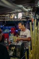 A man is standing at a street market stall, smiling as he operates a machine used for extracting sugarcane juice. Around him are several stalks of sugarcane and some plastic bottles. The scene is lit by overhead lights and features various red plastic stools in the background.