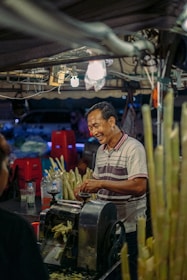 A man is standing at a street market stall, smiling as he operates a machine used for extracting sugarcane juice. Around him are several stalks of sugarcane and some plastic bottles. The scene is lit by overhead lights and features various red plastic stools in the background.