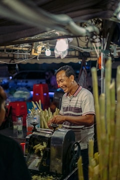 A man is standing at a street market stall, smiling as he operates a machine used for extracting sugarcane juice. Around him are several stalks of sugarcane and some plastic bottles. The scene is lit by overhead lights and features various red plastic stools in the background.