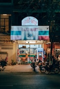 A small convenience store named 'Daily-Mart' is open at night. The store front is brightly lit with a sign displaying '24H'. Several people are gathered outside, including a woman using a vending machine and others on motorcycles parked nearby. The atmosphere suggests a casual and relaxed ambiance typical of a neighborhood store.