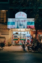 A small convenience store named 'Daily-Mart' is open at night. The store front is brightly lit with a sign displaying '24H'. Several people are gathered outside, including a woman using a vending machine and others on motorcycles parked nearby. The atmosphere suggests a casual and relaxed ambiance typical of a neighborhood store.
