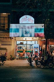 A small convenience store named 'Daily-Mart' is open at night. The store front is brightly lit with a sign displaying '24H'. Several people are gathered outside, including a woman using a vending machine and others on motorcycles parked nearby. The atmosphere suggests a casual and relaxed ambiance typical of a neighborhood store.