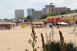 A sandy beach with several buildings in the background, including high-rise apartments and smaller structures with colorful decorations and red curtains. There are beach kiosks and signs like 'WC' indicating restrooms. A few people are walking along the beach, and plants are visible in the foreground.