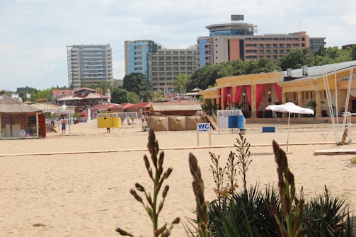 A sandy beach with several buildings in the background, including high-rise apartments and smaller structures with colorful decorations and red curtains. There are beach kiosks and signs like 'WC' indicating restrooms. A few people are walking along the beach, and plants are visible in the foreground.
