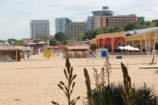 A sandy beach with several buildings in the background, including high-rise apartments and smaller structures with colorful decorations and red curtains. There are beach kiosks and signs like 'WC' indicating restrooms. A few people are walking along the beach, and plants are visible in the foreground.