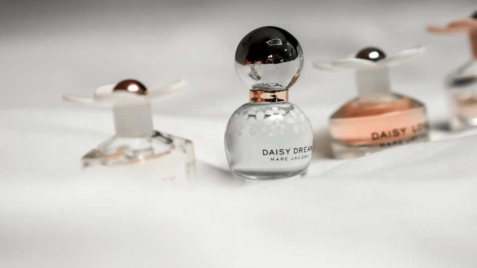 The image presents a collection of four perfume bottles, each with its own unique design and color. The first bottle is white with a gold cap and a silver flower on top, while the second bottle is pink with a gold cap and a silver flower on top. The third bottle is also white but has a black cap and a silver flower on top. The fourth bottle is brown with a gold cap and a silver flower on top. All four bottles are placed in a row against a gray background, creating an aesthetically pleasing composition that highlights the individual beauty of each perfume bottle.