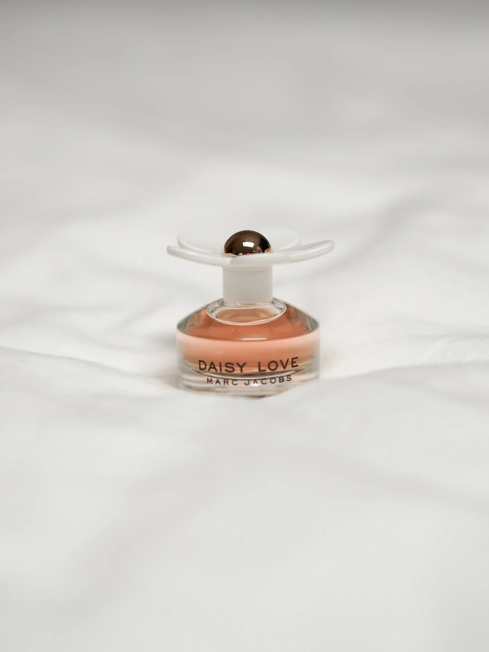 Small bottle of perfume Daisy Love sits on a white bedspread, partially to left