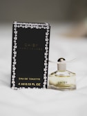 A small bottle of Daisy by Marc Jacobs perfume next to its black packaging. The bottle has a white flower-shaped cap with gold detailing. The packaging features small daisy designs around the edges.