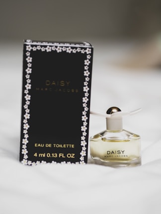 A small bottle of Daisy by Marc Jacobs perfume next to its black packaging. The bottle has a white flower-shaped cap with gold detailing. The packaging features small daisy designs around the edges.
