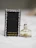 A small bottle of Daisy by Marc Jacobs perfume next to its black packaging. The bottle has a white flower-shaped cap with gold detailing. The packaging features small daisy designs around the edges.