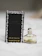 A small bottle of Daisy by Marc Jacobs perfume next to its black packaging. The bottle has a white flower-shaped cap with gold detailing. The packaging features small daisy designs around the edges.