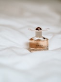 A perfume bottle with the label 'Daisy Marc Jacobs' sits on a soft, white bedspread. The bottle is elegant, with a transparent body and a white cap adorned with a golden top.