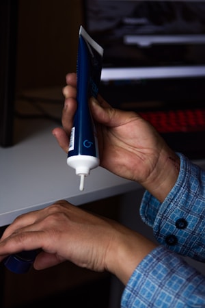 A person is holding a tube of cream, squeezing its contents onto the back of their other hand. The tube is blue with white accents and there is a computer or laptop keyboard in the background. They appear to be wearing a blue checkered shirt.