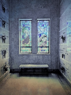 A solemn interior space with two stained glass windows depicting religious figures, possibly angels, casting colorful blue and purple hues. The stone walls are adorned with memorial plaques and small flower arrangements. A cushioned bench sits below the windows, enhancing the serene and reflective environment.