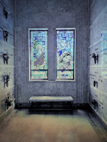 A solemn interior space with two stained glass windows depicting religious figures, possibly angels, casting colorful blue and purple hues. The stone walls are adorned with memorial plaques and small flower arrangements. A cushioned bench sits below the windows, enhancing the serene and reflective environment.