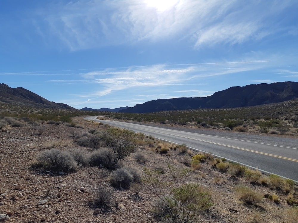 gray asphalt road near green grass field under blue and white sunny cloudy sky during daytime