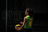 A person wearing a green and yellow sports jersey with the name and number 'Jurado 10' is standing outdoors holding a blue and yellow volleyball. The setting appears to be shadowy, possibly a sports court or outdoor area with railings visible in the background.