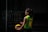 A person wearing a green and yellow sports jersey with the name and number 'Jurado 10' is standing outdoors holding a blue and yellow volleyball. The setting appears to be shadowy, possibly a sports court or outdoor area with railings visible in the background.