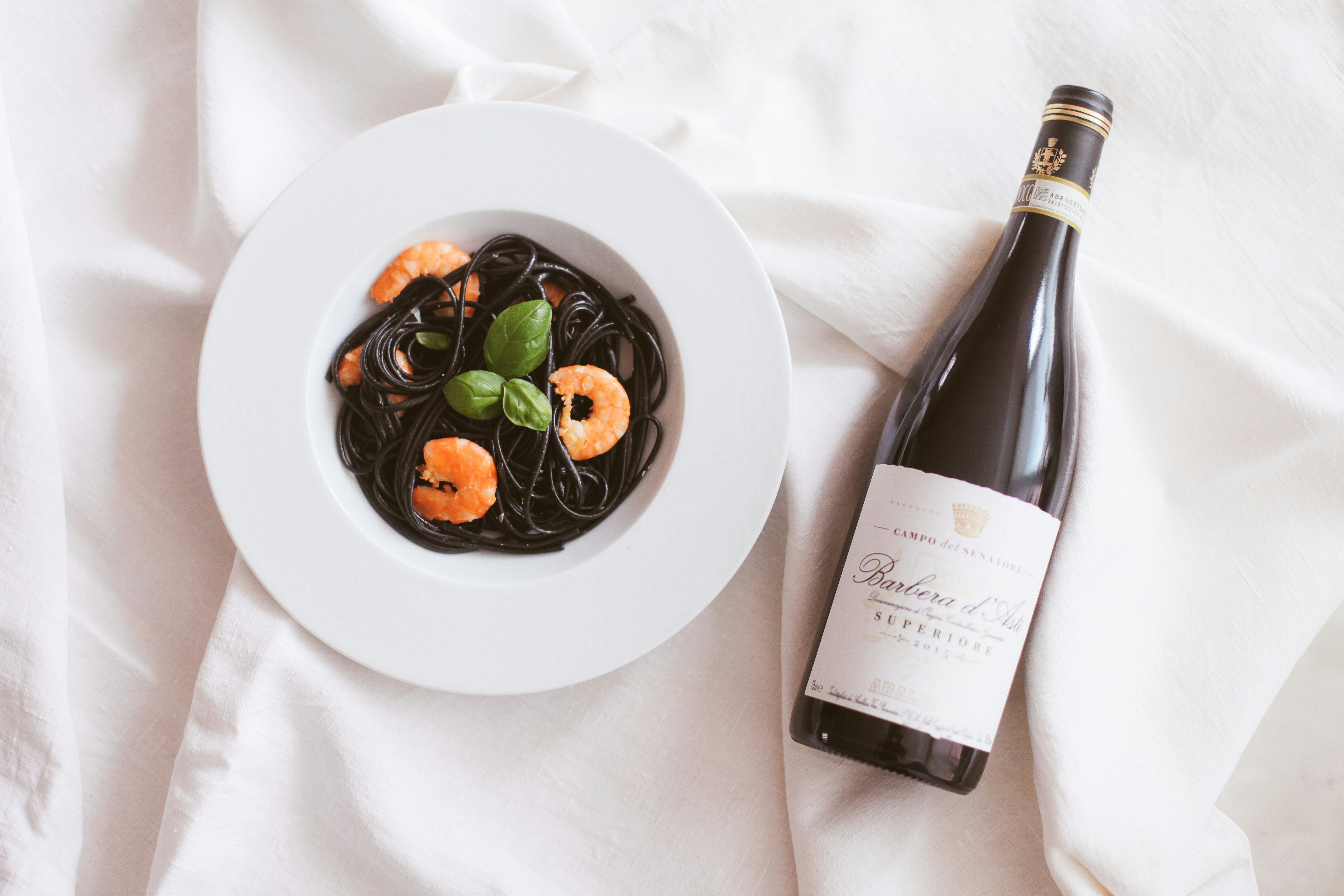 white ceramic plate with green vegetable and wine bottle, A bottle of red wine and black past with shrimps and basil on white tableware
