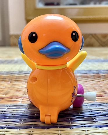 A brightly colored plastic toy duck with a round body. It is predominantly orange with a blue beak and blue accents on the sides. The wings are yellow, and there is a wind-up key attached to its side. The duck is placed on a woven surface with a mix of purple and natural colors.
