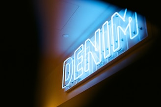 A bright blue neon sign with the word 'DENIM' is illuminated against a darker background. The sign is mounted on a wall, with screws visible at its base, and emits a cool and vibrant light that casts a slight glow around it. The surroundings are dimly lit, causing the sign to stand out prominently.