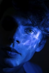A close-up portrait of a person partially illuminated by blue light, creating a mysterious and artistic effect. The lighting casts intricate patterns across the face, highlighting the eyes and lips while leaving parts of the face in shadow.