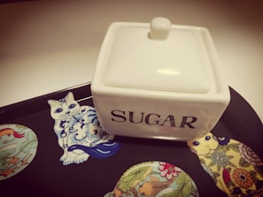 A white, square ceramic sugar container with a lid is placed on a decorative tray. The tray features colorful illustrations of stylized animals, including a cat and birds, with intricate patterns. The word 'SUGAR' is prominently displayed on the container in bold letters.