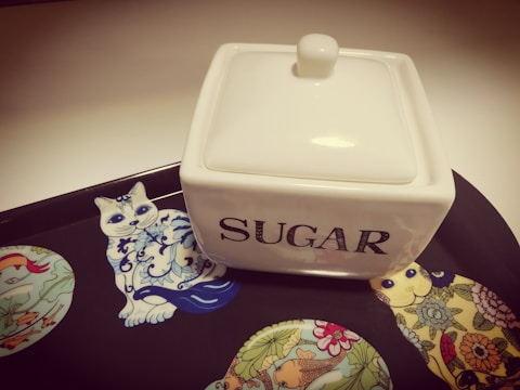 A white, square ceramic sugar container with a lid is placed on a decorative tray. The tray features colorful illustrations of stylized animals, including a cat and birds, with intricate patterns. The word 'SUGAR' is prominently displayed on the container in bold letters.