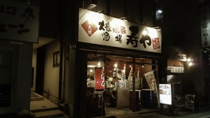 A warmly lit restaurant entrance with a bright sign in Japanese characters. Inside, several patrons are seated, engaged in dining, while the exterior has traditional lanterns and banners adding to the ambiance.