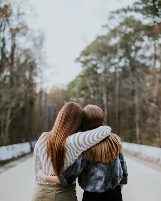 Two people with long hair embracing on a road lined with tall trees