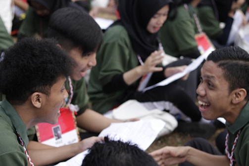 A group of young people are sitting together, engaged in lively conversation and possibly studying or participating in a discussion. They are wearing green shirts and have name tags. Some are holding papers, and they all seem to be enjoying the moment with smiles and animated expressions.