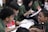 A group of young people are sitting together, engaged in lively conversation and possibly studying or participating in a discussion. They are wearing green shirts and have name tags. Some are holding papers, and they all seem to be enjoying the moment with smiles and animated expressions.