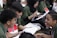 A group of young people are sitting together, engaged in lively conversation and possibly studying or participating in a discussion. They are wearing green shirts and have name tags. Some are holding papers, and they all seem to be enjoying the moment with smiles and animated expressions.