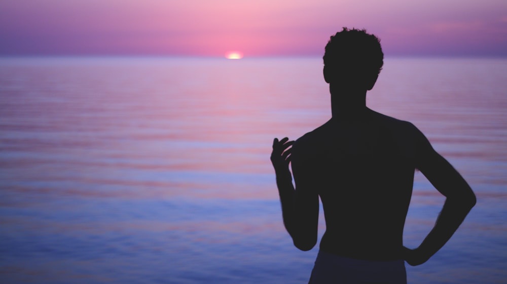 silhouette of man standing near body of water during sunset